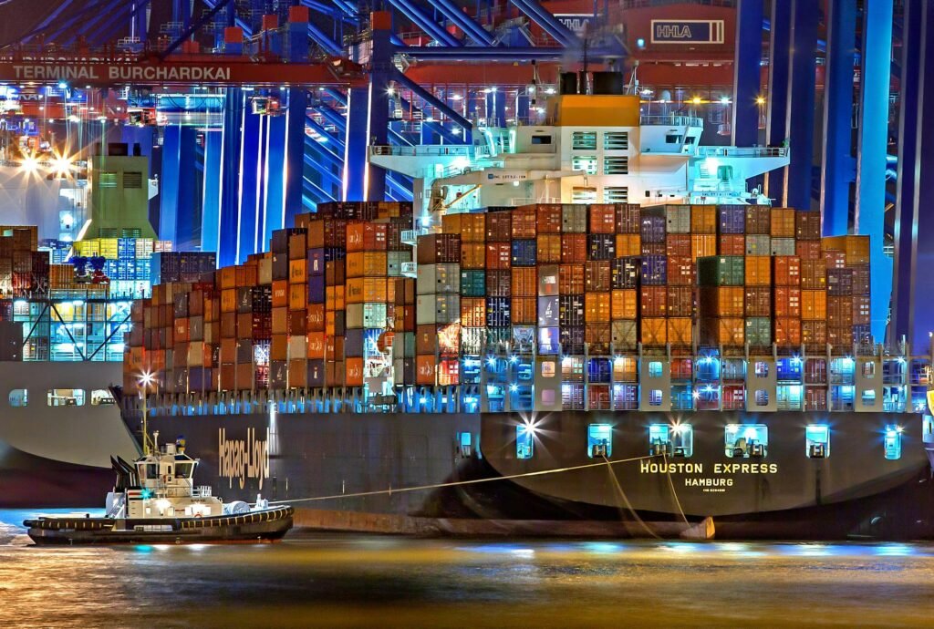 pexels-photo-753331-753331-1 A brightly lit cargo ship at Hamburg harbor with stacked containers and a tugboat.